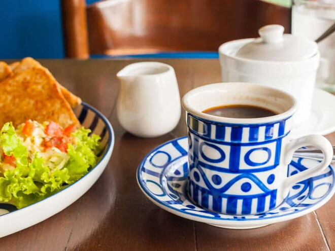 Una taza blanca y azul que contiene café y al lado un plato con ensalada verde y quesadillas